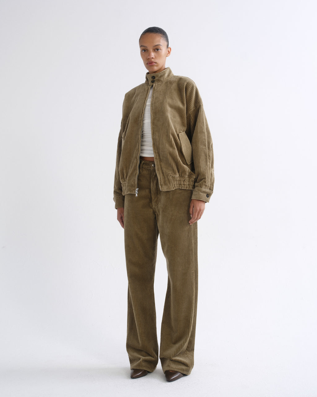 A model stands against a plain white studio background wearing a matching corduroy jacket and trousers set in a muted olive-brown color, styled with a white ribbed top and brown shoes.