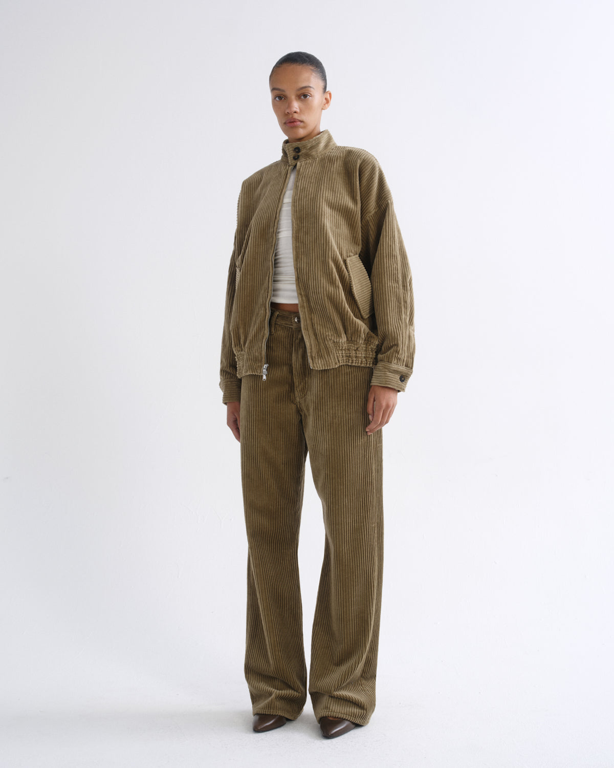 A model stands against a plain white studio background wearing a matching corduroy jacket and trousers set in a muted olive-brown color, styled with a white ribbed top and brown shoes.