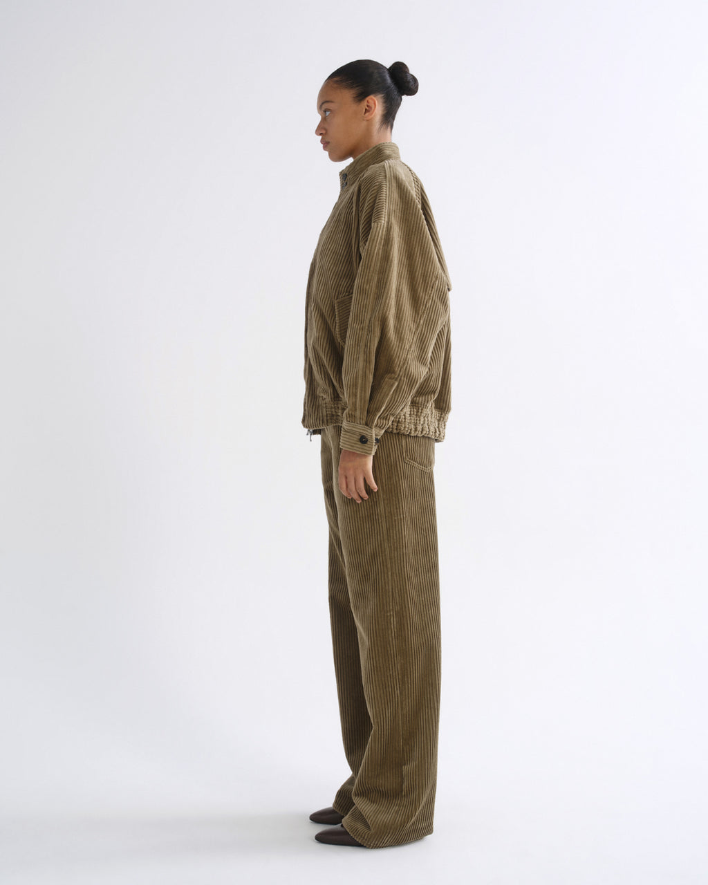A model stands in profile wearing a matching corduroy jacket and wide-leg trousers set in a muted olive-brown color, photographed against a seamless light gray studio background.