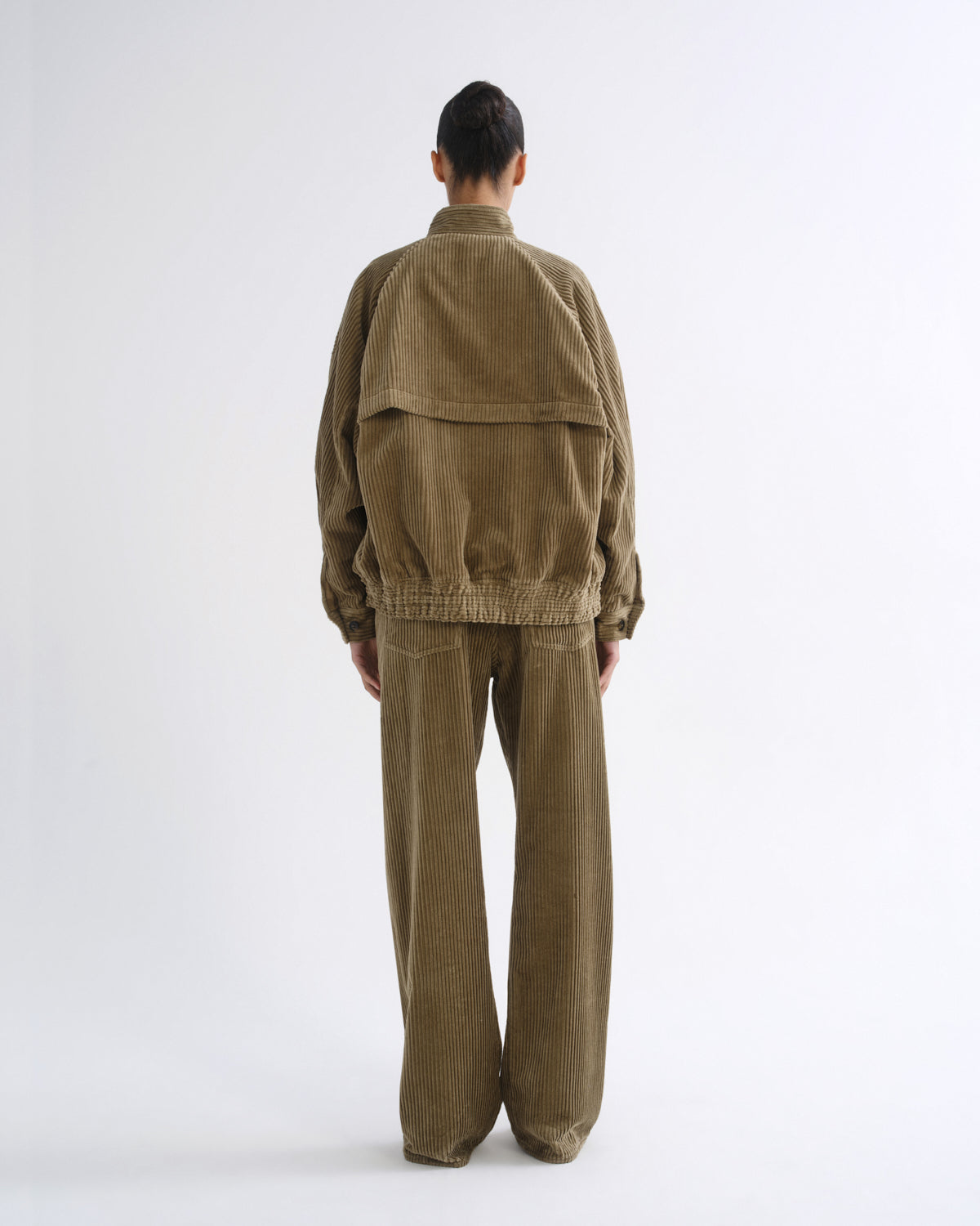 A model viewed from the back wearing an olive brown corduroy jacket and matching wide-leg trousers against a plain white studio background.