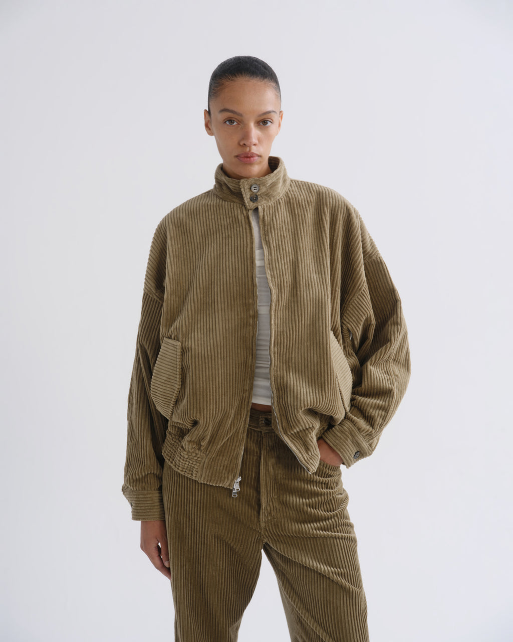 A model wearing a brown corduroy jacket and matching corduroy pants over a striped top, photographed against a plain light gray studio background.