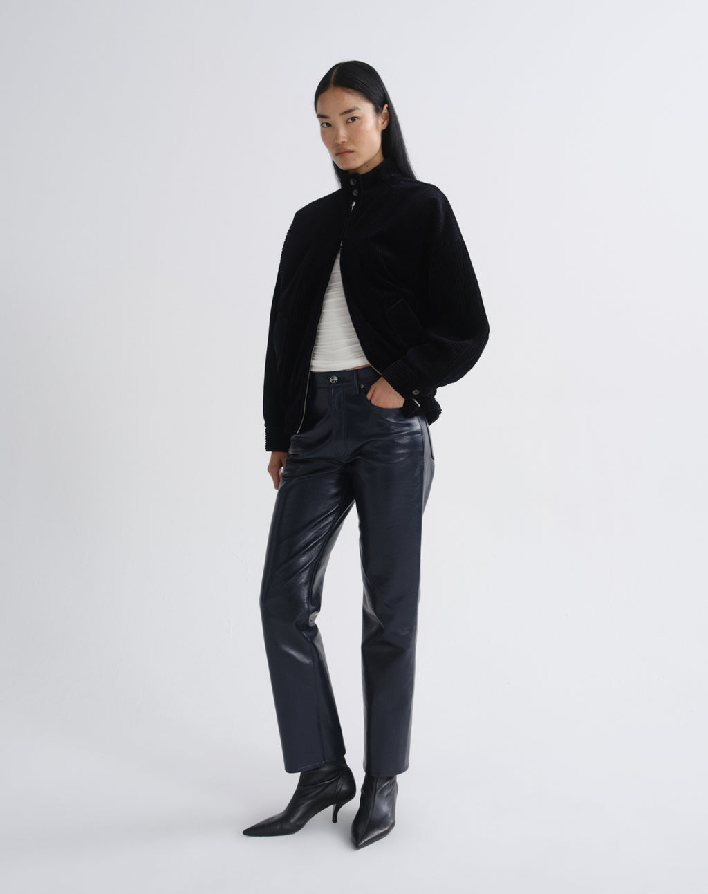 Person wearing a black bomber jacket with dark grey straight leg leather pants viewed from the side on a grey background