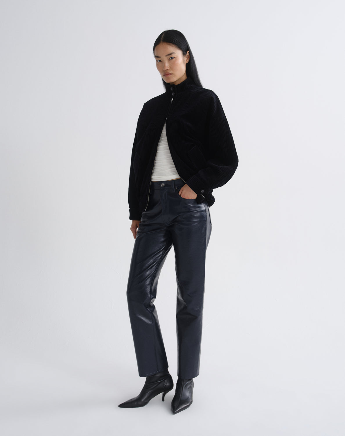 Person wearing a black bomber jacket with dark grey straight leg leather pants viewed from the side on a grey background