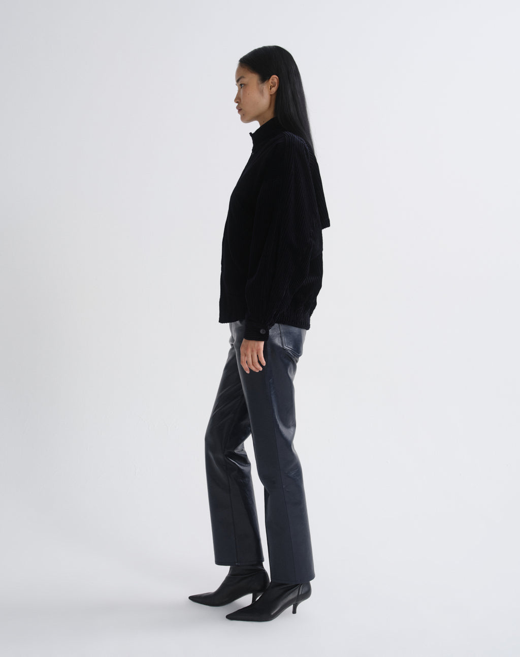 Person wearing a black bomber jacket with dark grey straight leg leather pants viewed from the side on a grey background