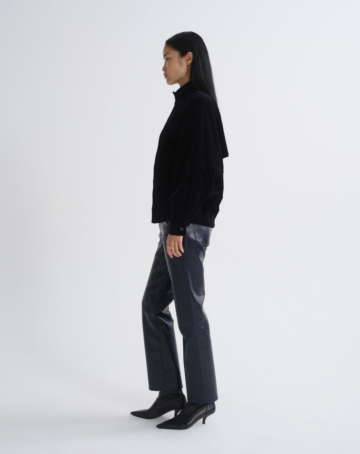 Person wearing a black bomber jacket with dark grey straight leg leather pants viewed from the side on a grey background