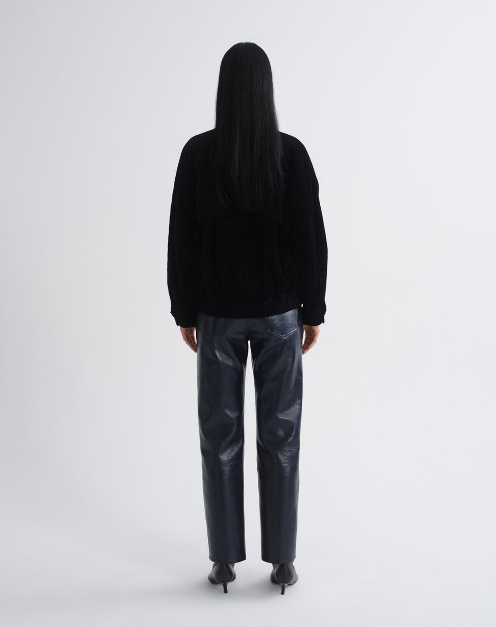 Person wearing a black bomber jacket with dark grey straight leg leather pants viewed from the back on a grey background