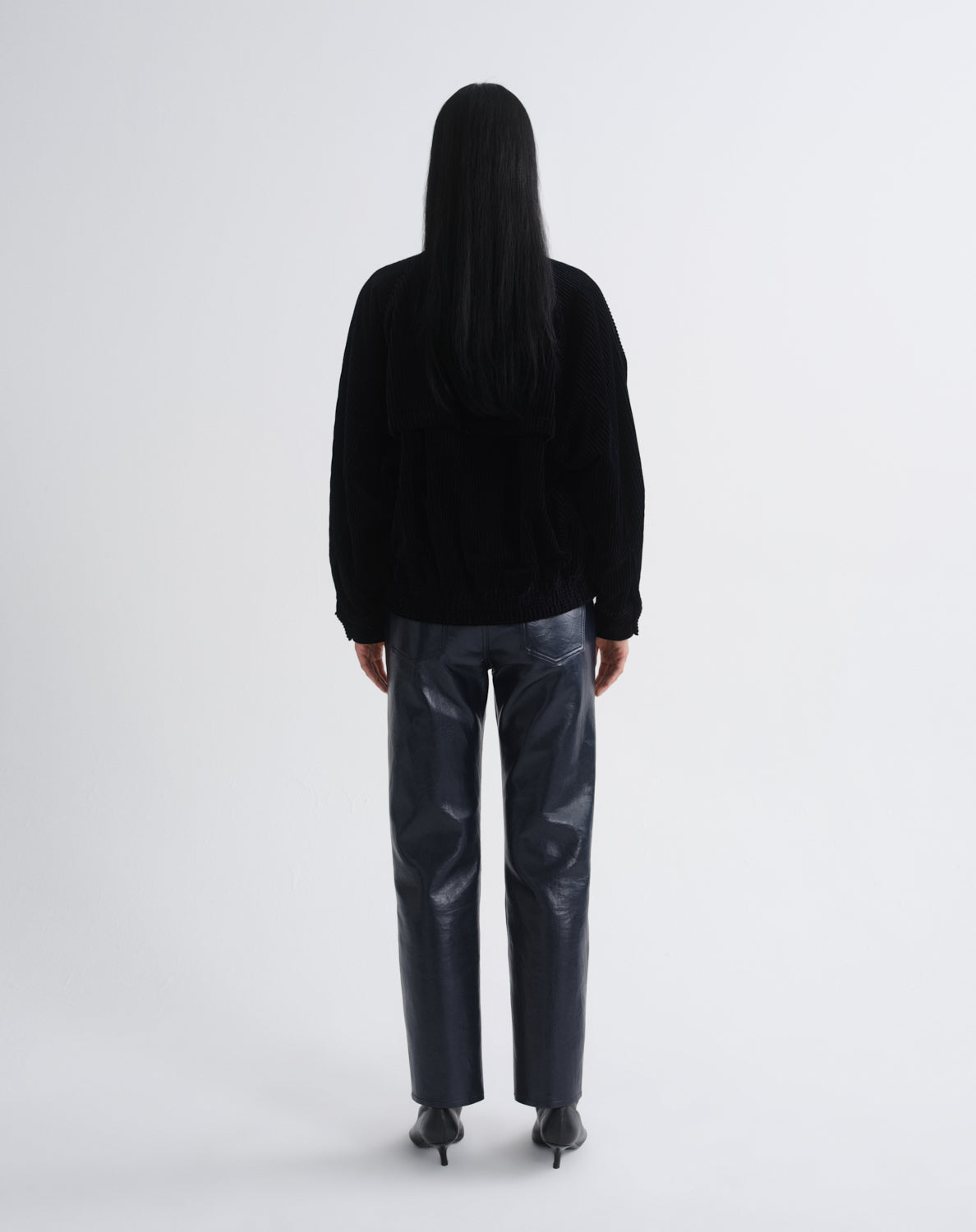 Person wearing a black bomber jacket with dark grey straight leg leather pants viewed from the back on a grey background