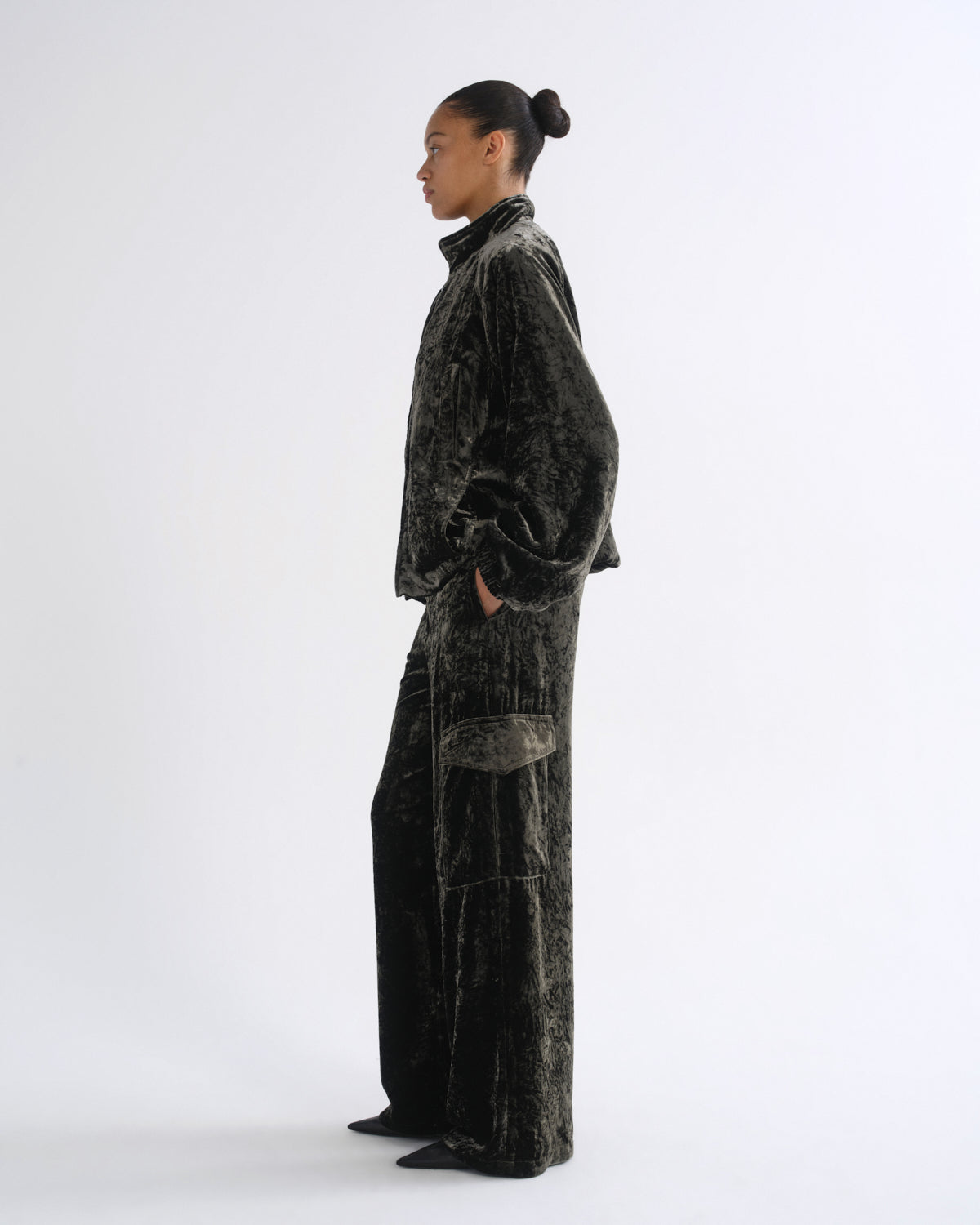 Woman wearing a black velvet long sleeve top with black velvet pants viewed from the side on a grey background.
