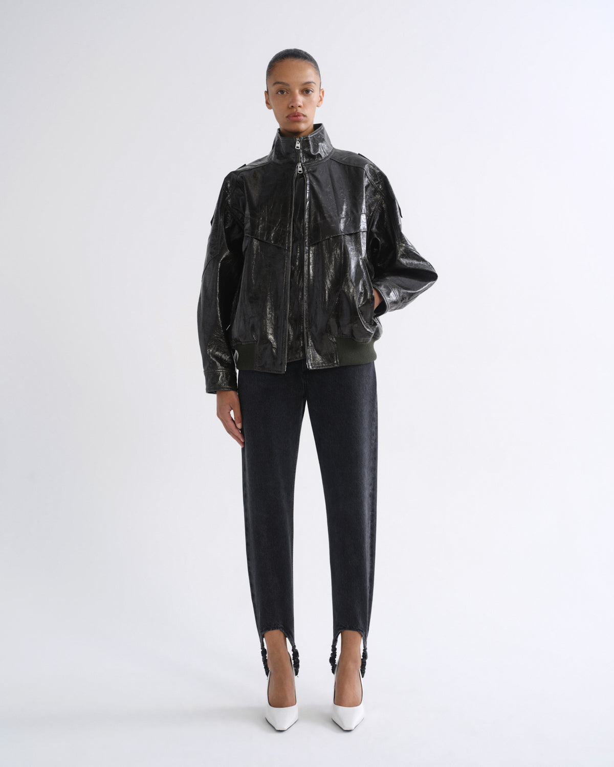 A model stands against a light gray studio background wearing a shiny black cropped jacket with a high collar, black cropped pants, and white pointed heels.