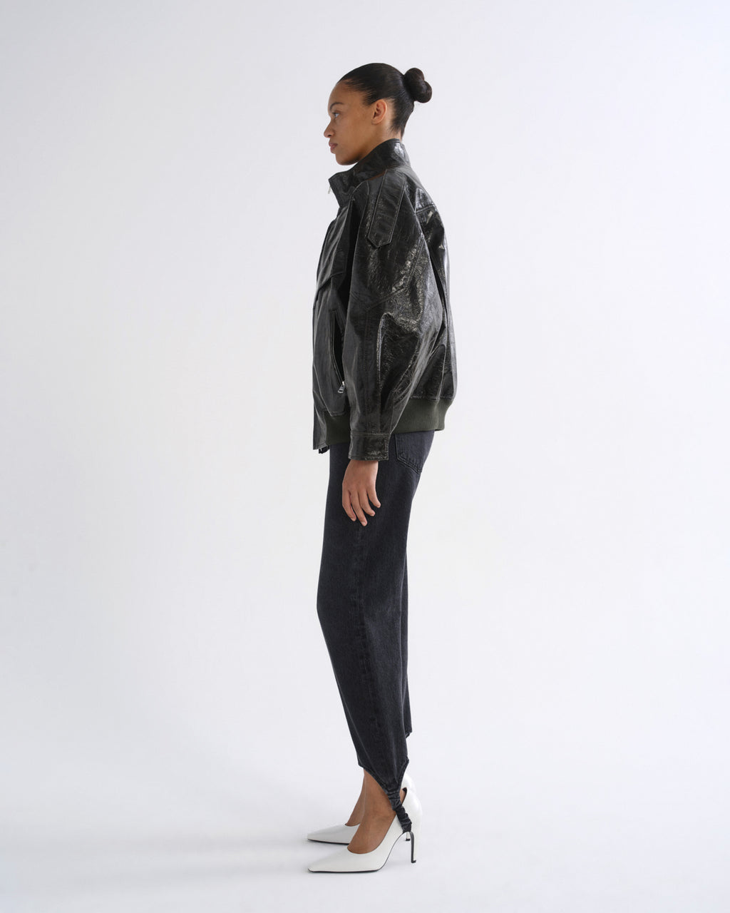 A model shown in profile wearing a black distressed leather jacket, dark charcoal pants, and white pointed-toe heels against a plain white studio background.