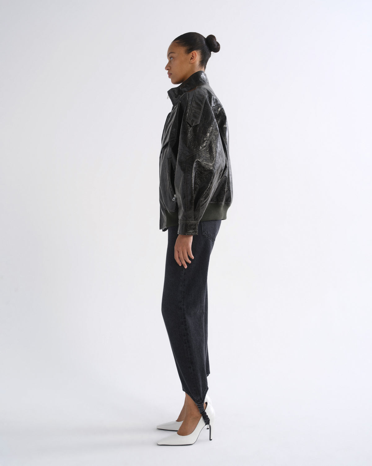 A model shown in profile wearing a black distressed leather jacket, dark charcoal pants, and white pointed-toe heels against a plain white studio background.