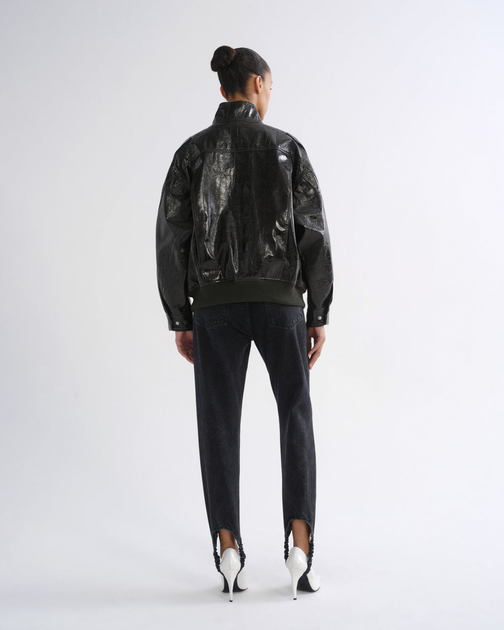 A model viewed from the back wearing a black leather jacket with a glossy, crumpled plastic-like texture on the back panels, paired with dark jeans and white high-heeled shoes, against a plain light gray studio background.