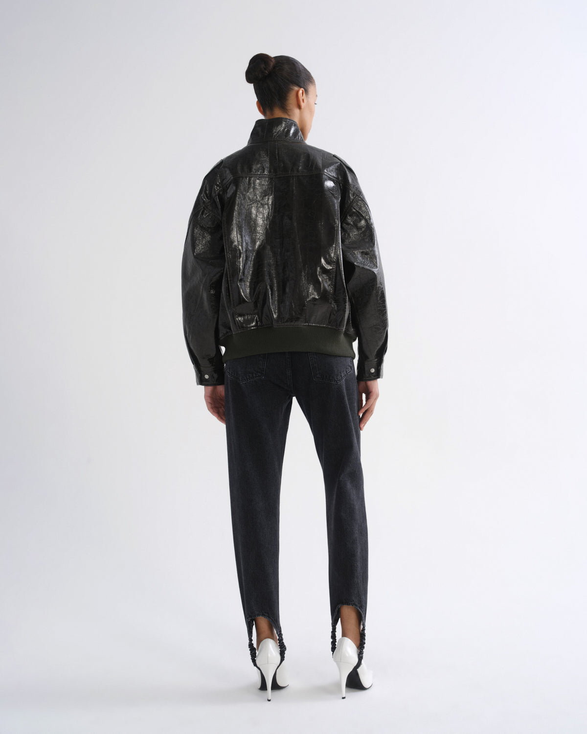 A model viewed from the back wearing a black leather jacket with a glossy, crumpled plastic-like texture on the back panels, paired with dark jeans and white high-heeled shoes, against a plain light gray studio background.
