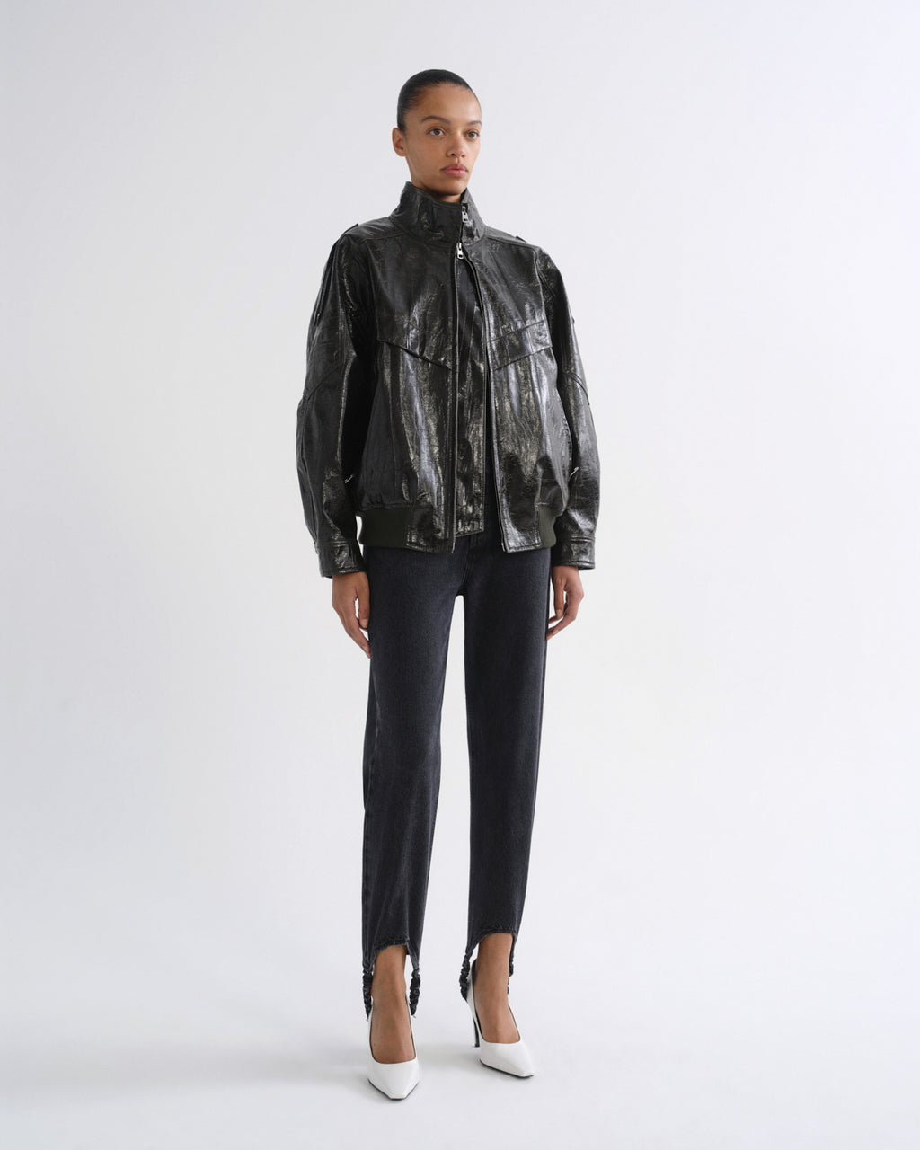 A model stands against a light gray studio background wearing a shiny black cropped jacket with a high collar, black cropped pants, and white pointed heels.