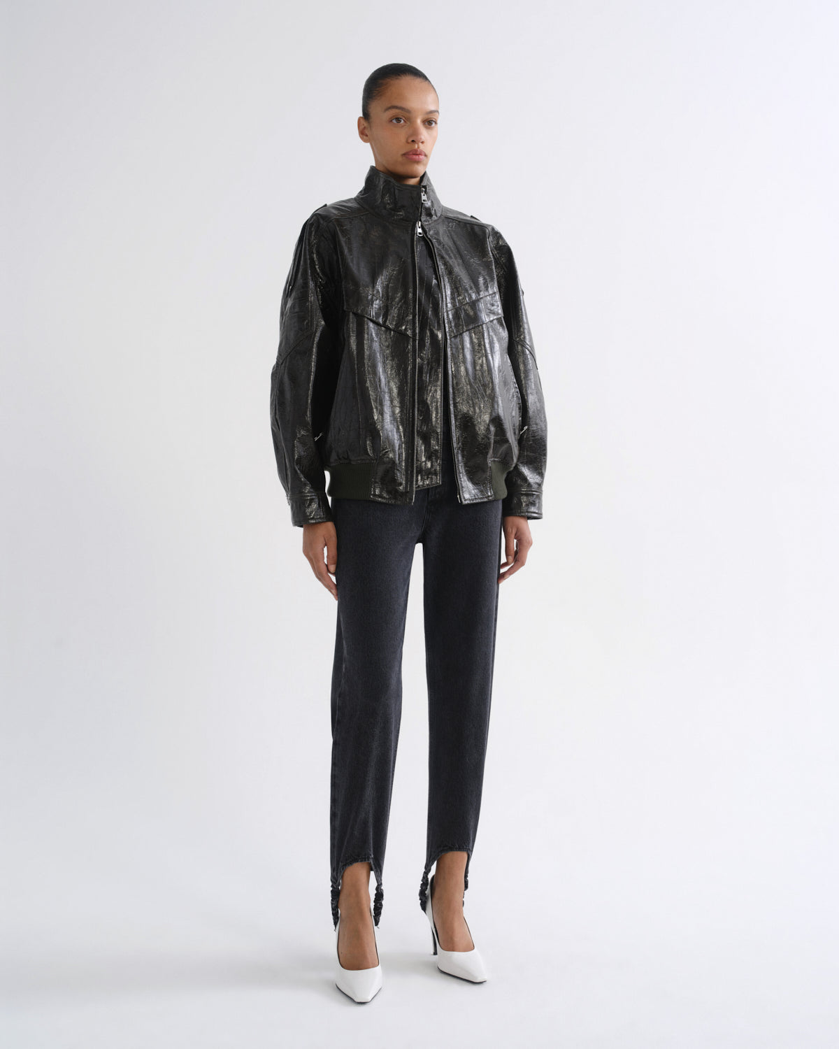 A model stands against a light gray studio background wearing a shiny black cropped jacket with a high collar, black cropped pants, and white pointed heels.