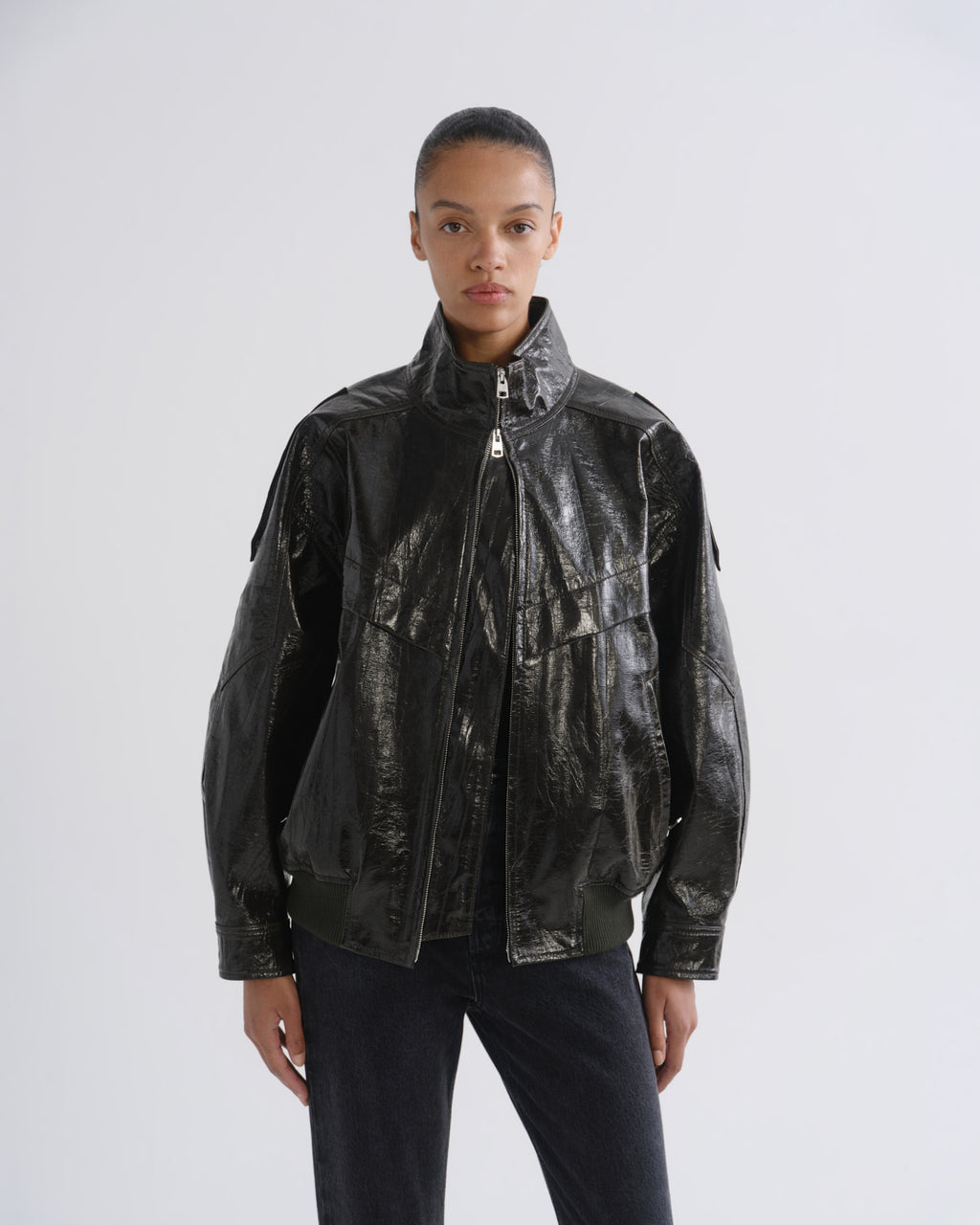 A model wearing a shiny black leather jacket with a high collar and front zipper, paired with dark pants, photographed against a light gray studio background.