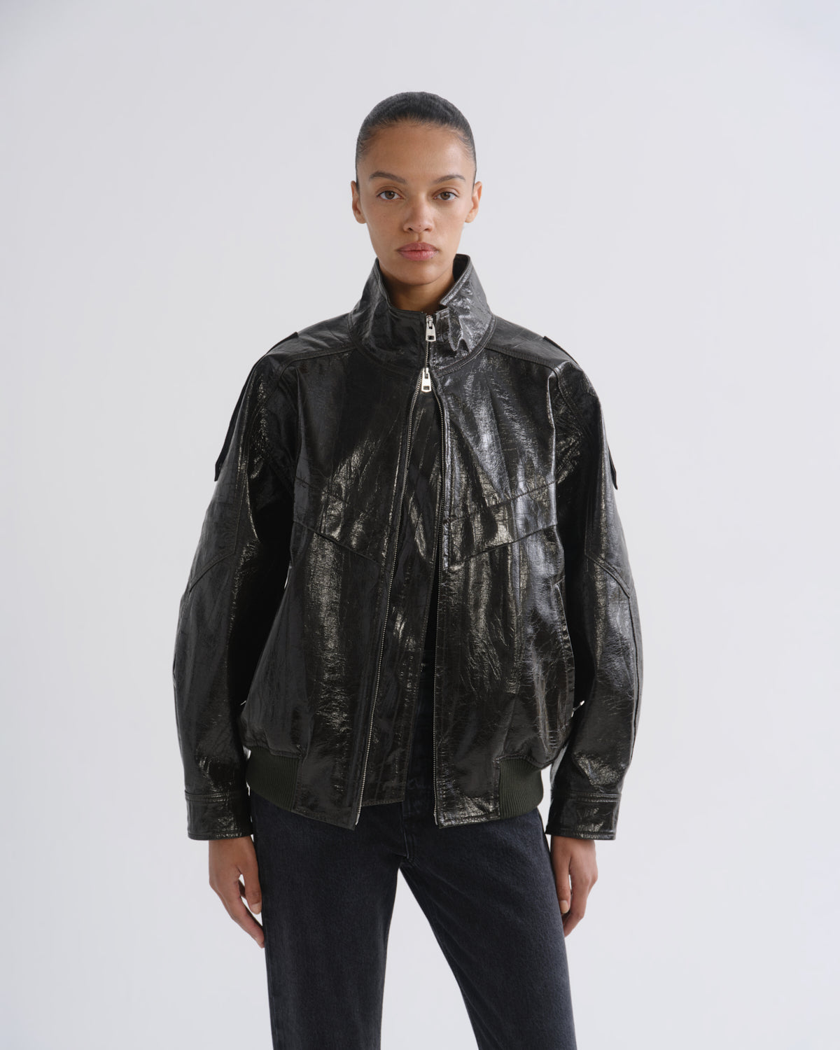 A model wearing a shiny black leather jacket with a high collar and front zipper, paired with dark pants, photographed against a light gray studio background.