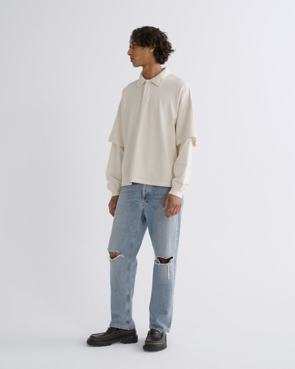 A model stands in profile wearing a cream-colored, long-sleeved collared shirt with layered sleeves, light-wash distressed blue jeans, and black leather shoes, against a plain light gray studio background.