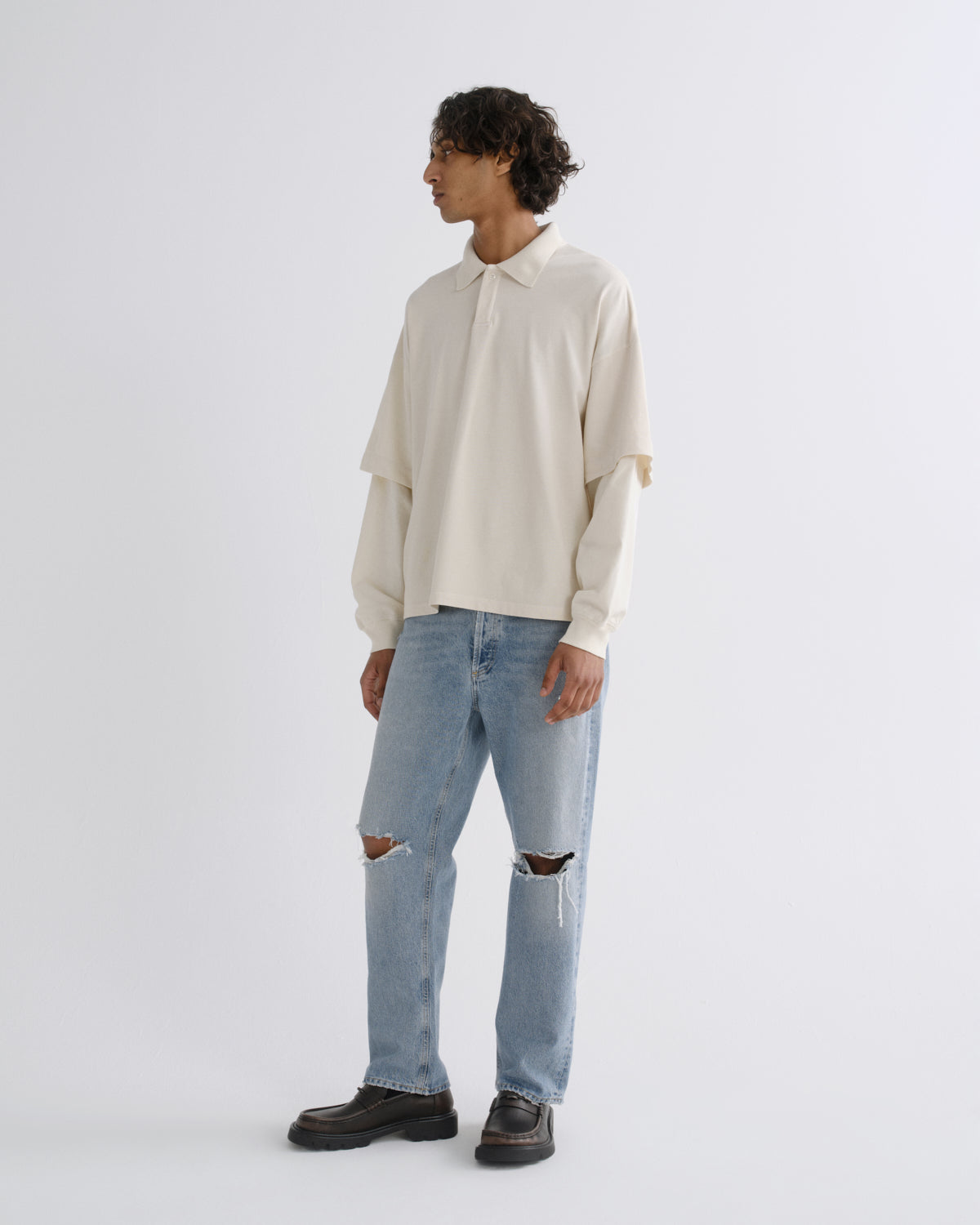 A model stands in profile wearing a cream-colored, long-sleeved collared shirt with layered sleeves, light-wash distressed blue jeans, and black leather shoes, against a plain light gray studio background.