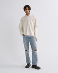 A model stands against a light gray studio background wearing a cream long-sleeve collared shirt, light-wash distressed jeans, and dark brown leather loafers.