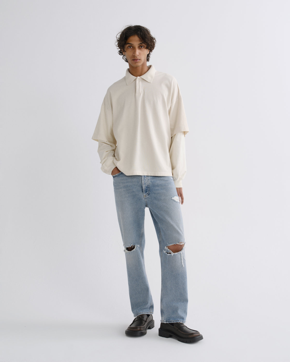 A model stands against a light gray studio background wearing a cream long-sleeve collared shirt, light-wash distressed jeans, and dark brown leather loafers.