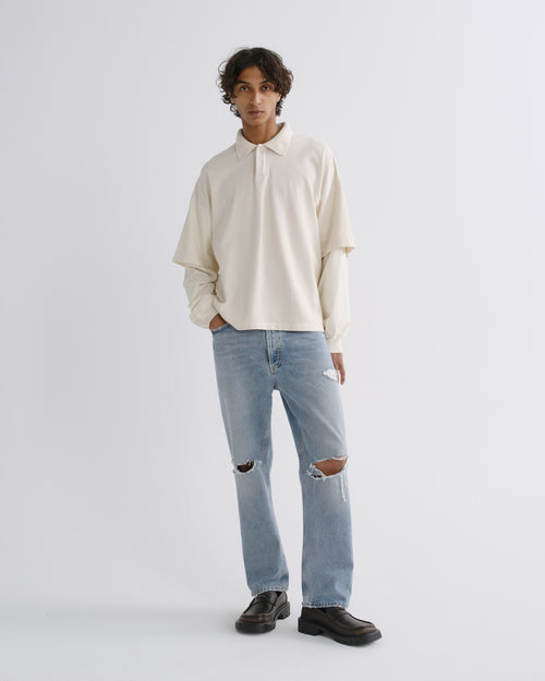 A model stands against a light gray studio background wearing a cream long-sleeve collared shirt, light-wash distressed jeans, and dark brown leather loafers.