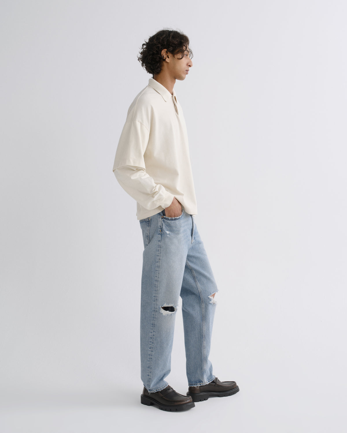 A man wearing a cream long-sleeve button-up shirt, light-wash ripped blue jeans, and dark brown leather shoes, standing in profile against a light gray studio background.