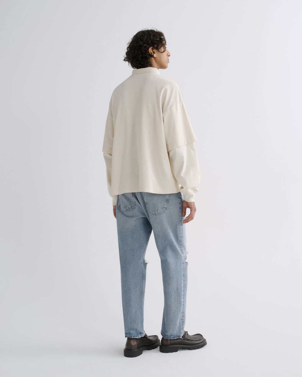 A man is shown from the back wearing a cream-colored long-sleeve shirt, light blue jeans, and dark brown loafers, standing against a plain light gray studio background.
