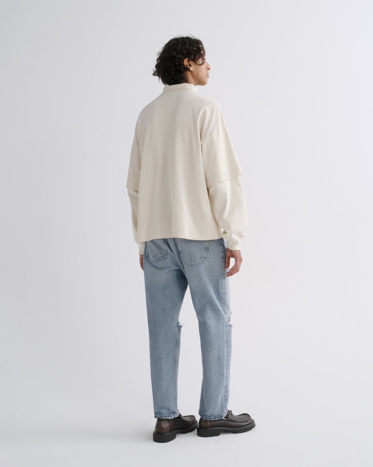 A man is shown from the back wearing a cream-colored long-sleeve shirt, light blue jeans, and dark brown loafers, standing against a plain light gray studio background.
