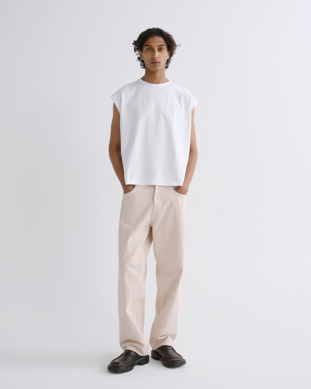 A model stands against a light gray studio background wearing a white sleeveless crewneck t-shirt, light beige straight-leg trousers, and dark brown leather loafers.