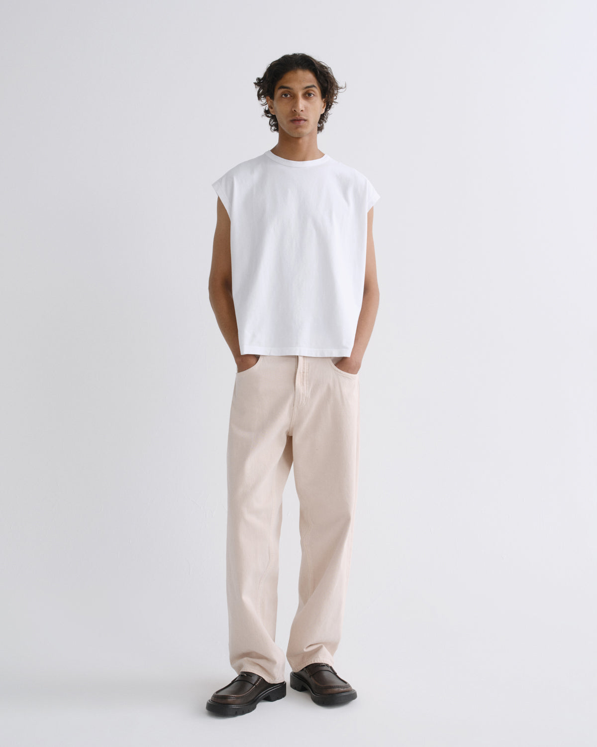 A model stands against a light gray studio background wearing a white sleeveless crewneck t-shirt, light beige straight-leg trousers, and dark brown leather loafers.