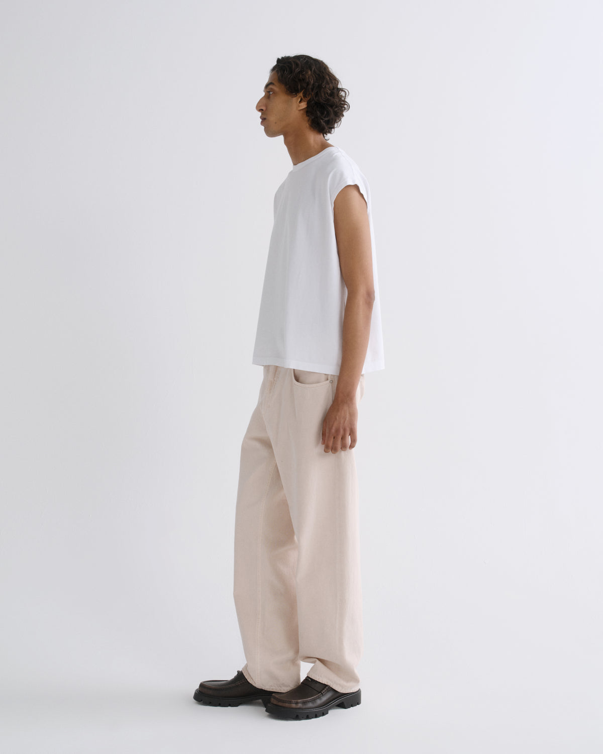 A model shown in profile wearing a white sleeveless t-shirt, beige wide-leg trousers, and dark brown leather loafers against a plain light gray studio background.