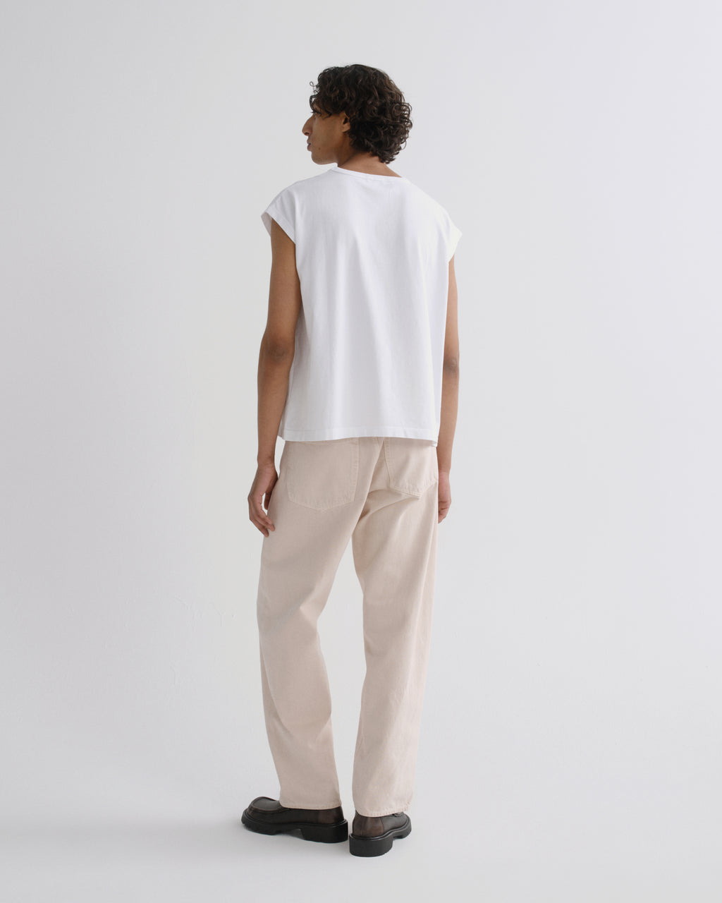 A model shown from the back wearing a white sleeveless t-shirt and beige trousers, standing against a plain light gray studio background.