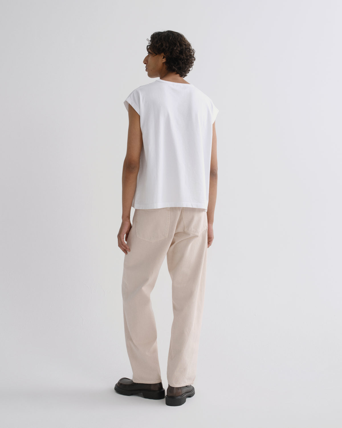 A model shown from the back wearing a white sleeveless t-shirt and beige trousers, standing against a plain light gray studio background.