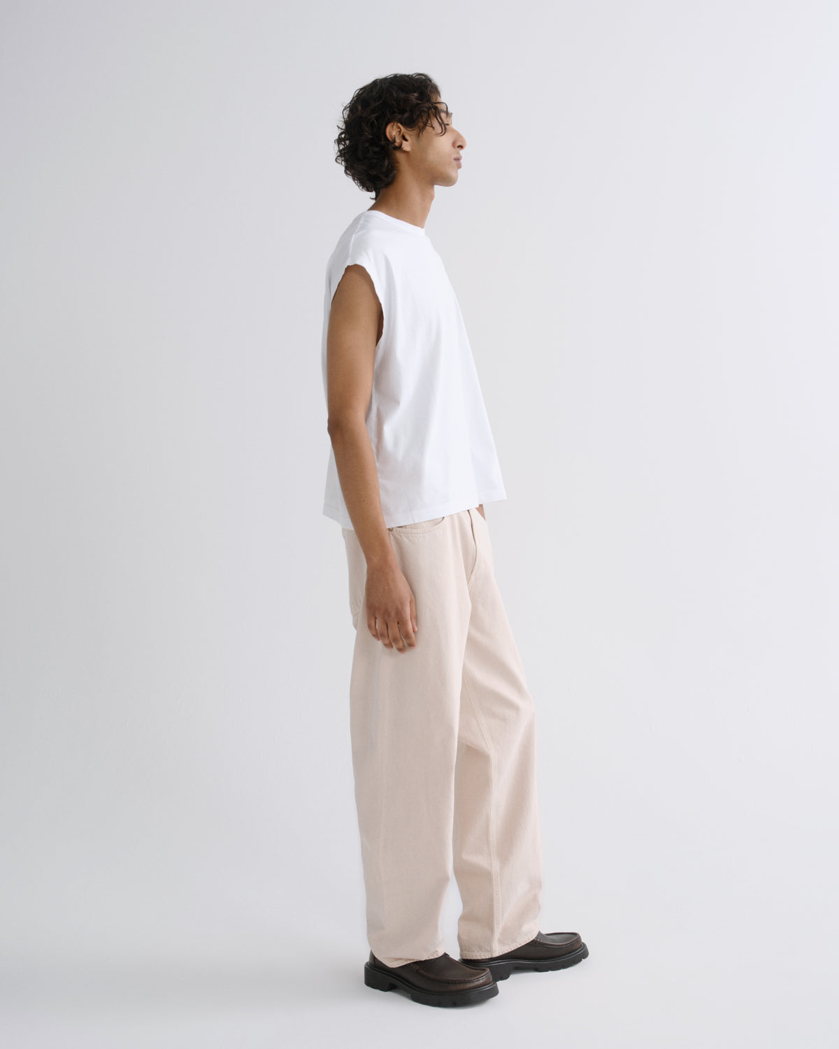 A man in profile wearing a white sleeveless t-shirt, light beige wide-leg trousers, and dark brown loafers, standing against a plain light gray studio background.