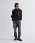 A man wearing a dark navy long-sleeve crewneck sweatshirt, blue jeans, and black leather shoes, standing against a light gray studio background.