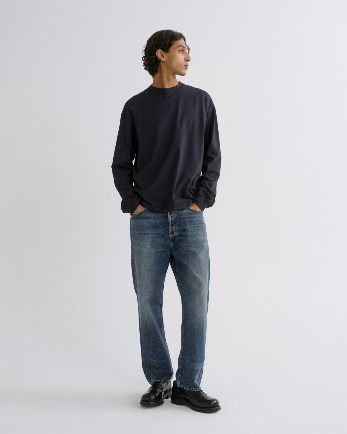 A man wearing a dark navy long-sleeve crewneck sweatshirt, blue jeans, and black leather shoes, standing against a light gray studio background.