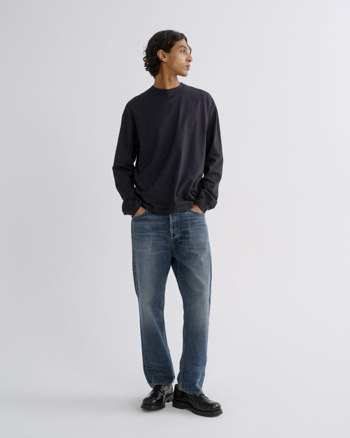 A man wearing a dark navy long-sleeve crewneck sweatshirt, blue jeans, and black leather shoes, standing against a light gray studio background.