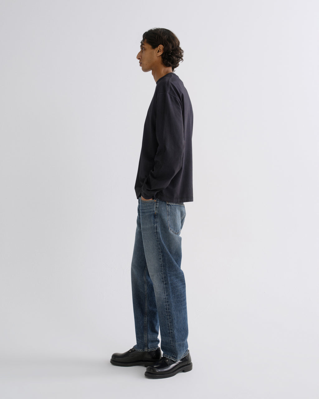 A man stands in profile wearing a dark navy long-sleeve shirt, blue jeans, and black leather shoes against a plain light gray studio background.