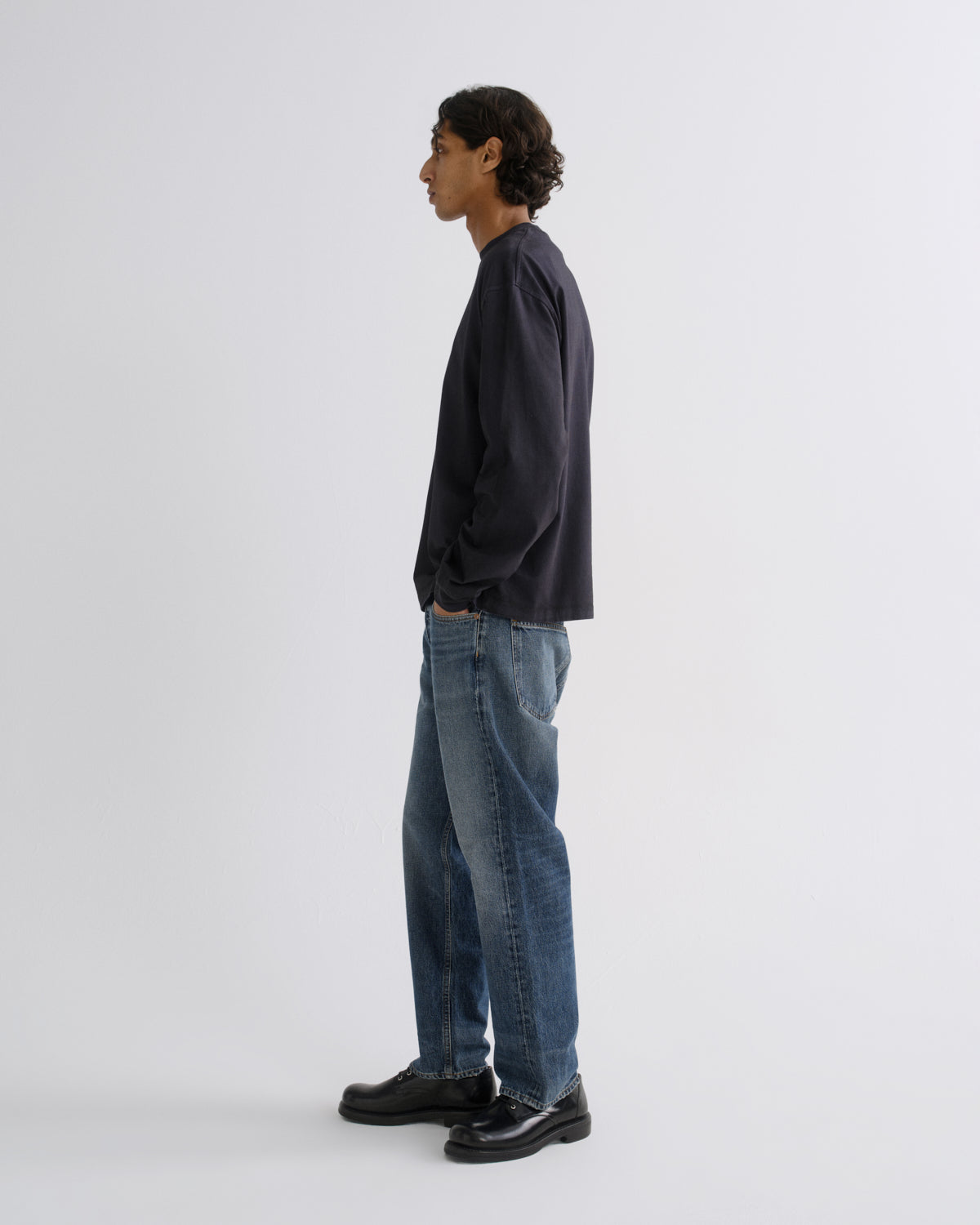A man stands in profile wearing a dark navy long-sleeve shirt, blue jeans, and black leather shoes against a plain light gray studio background.