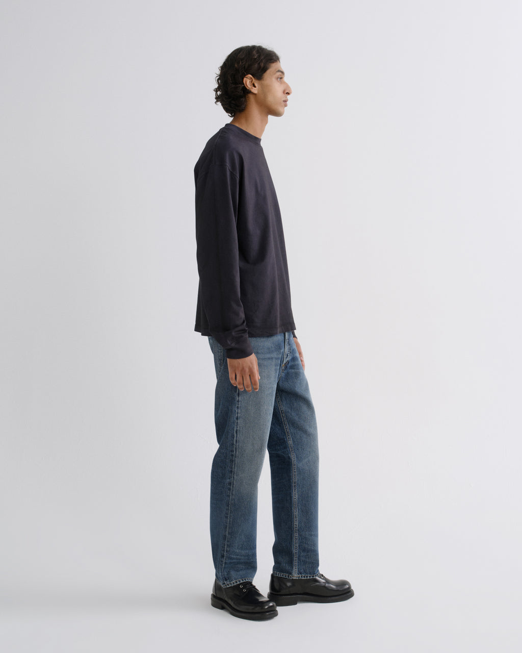 A man shown in profile wearing a dark navy long-sleeve crewneck sweatshirt, blue jeans, and black leather shoes, standing against a light gray studio background.