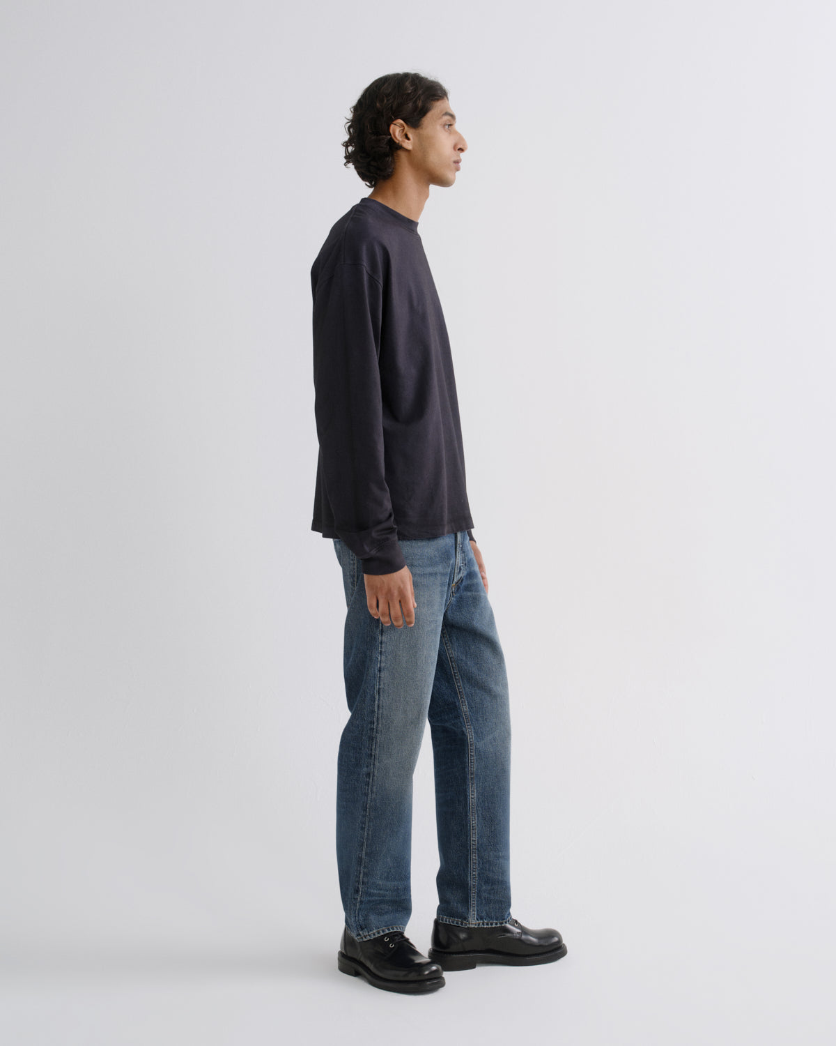 A man shown in profile wearing a dark navy long-sleeve crewneck sweatshirt, blue jeans, and black leather shoes, standing against a light gray studio background.