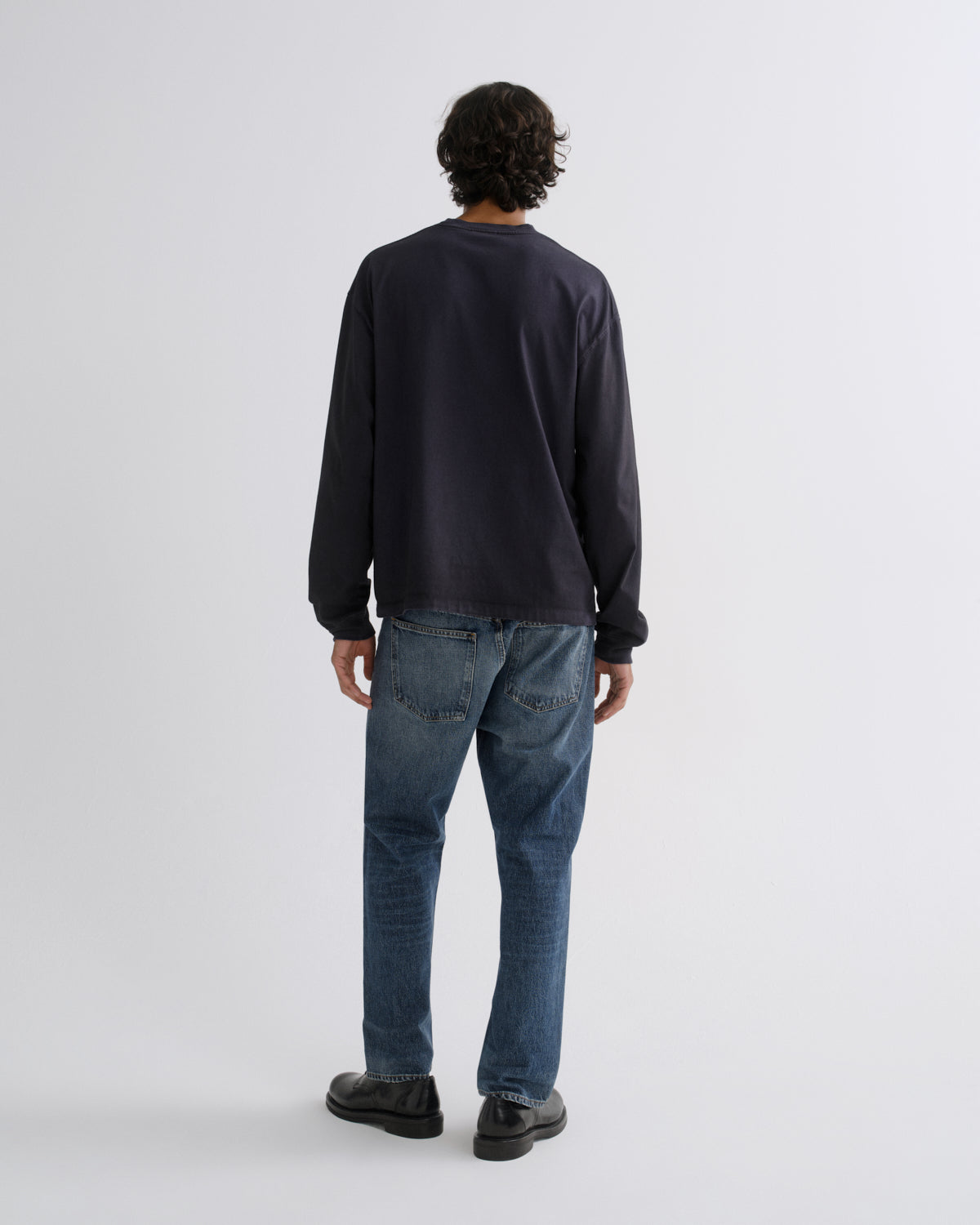 A man viewed from behind wearing a dark navy long-sleeve crewneck sweatshirt, blue jeans, and black shoes, standing against a light gray studio background.