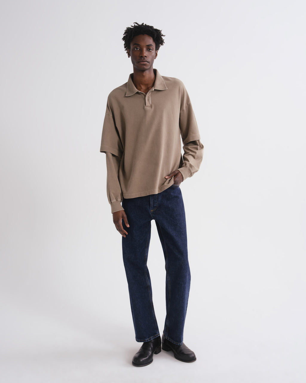 A man modeling a beige long-sleeve polo shirt with dark blue jeans and black shoes, standing against a plain white studio background.