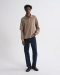 A man modeling a beige long-sleeve polo shirt with dark blue jeans and black shoes, standing against a plain white studio background.