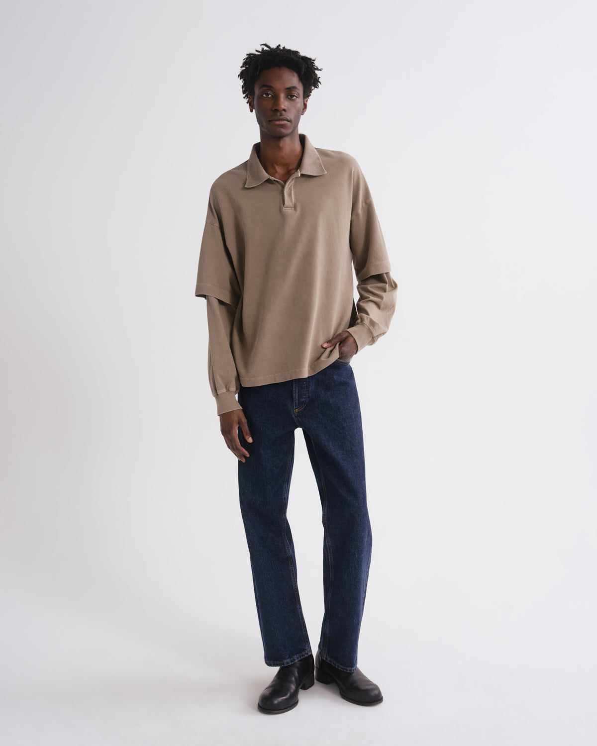 A man modeling a beige long-sleeve polo shirt with dark blue jeans and black shoes, standing against a plain white studio background.