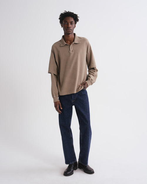 A man modeling a beige long-sleeve polo shirt with dark blue jeans and black shoes, standing against a plain white studio background.