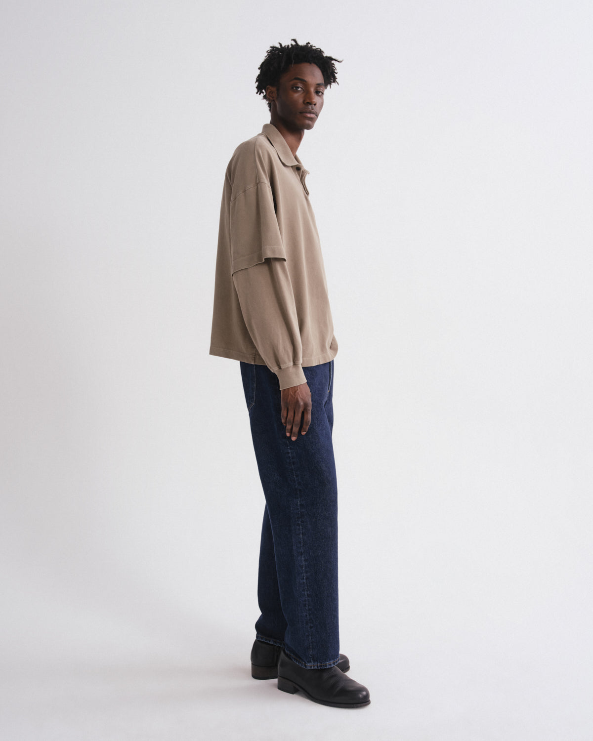 A man stands in profile wearing a beige oversized short-sleeve button-up shirt, dark blue jeans, and black shoes against a plain light gray studio background.