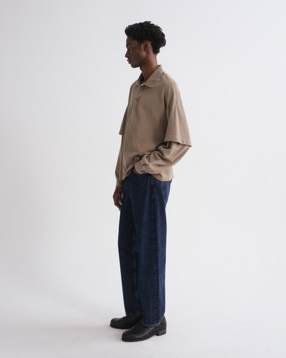 A man stands in profile wearing a beige long-sleeve collared shirt, dark blue wide-leg jeans, and black leather shoes against a plain light gray studio background.