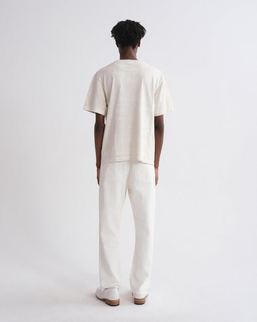 A model viewed from behind wearing a cream-colored short-sleeve t-shirt and matching cream-colored trousers, standing against a plain white studio background.